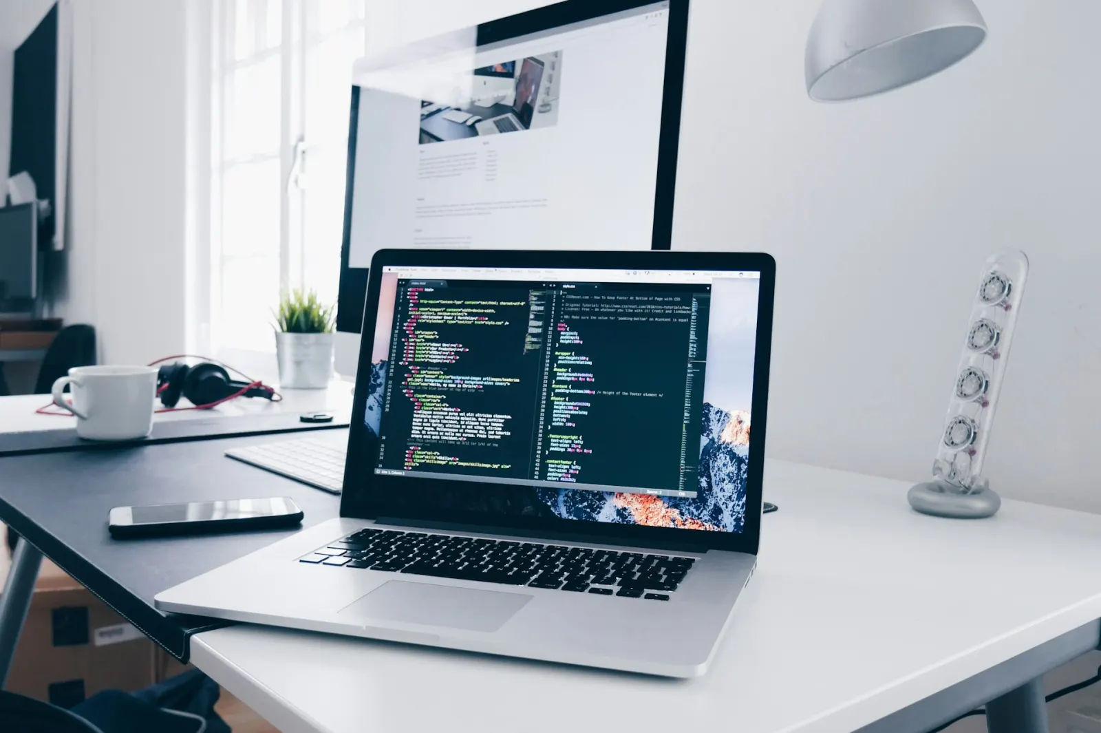 Schreibtisch mit Laptop, Monitor und Code-Editor im Tageslicht